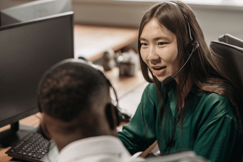 A smiling support agent wearing a headset works at a desk beside a colleague.